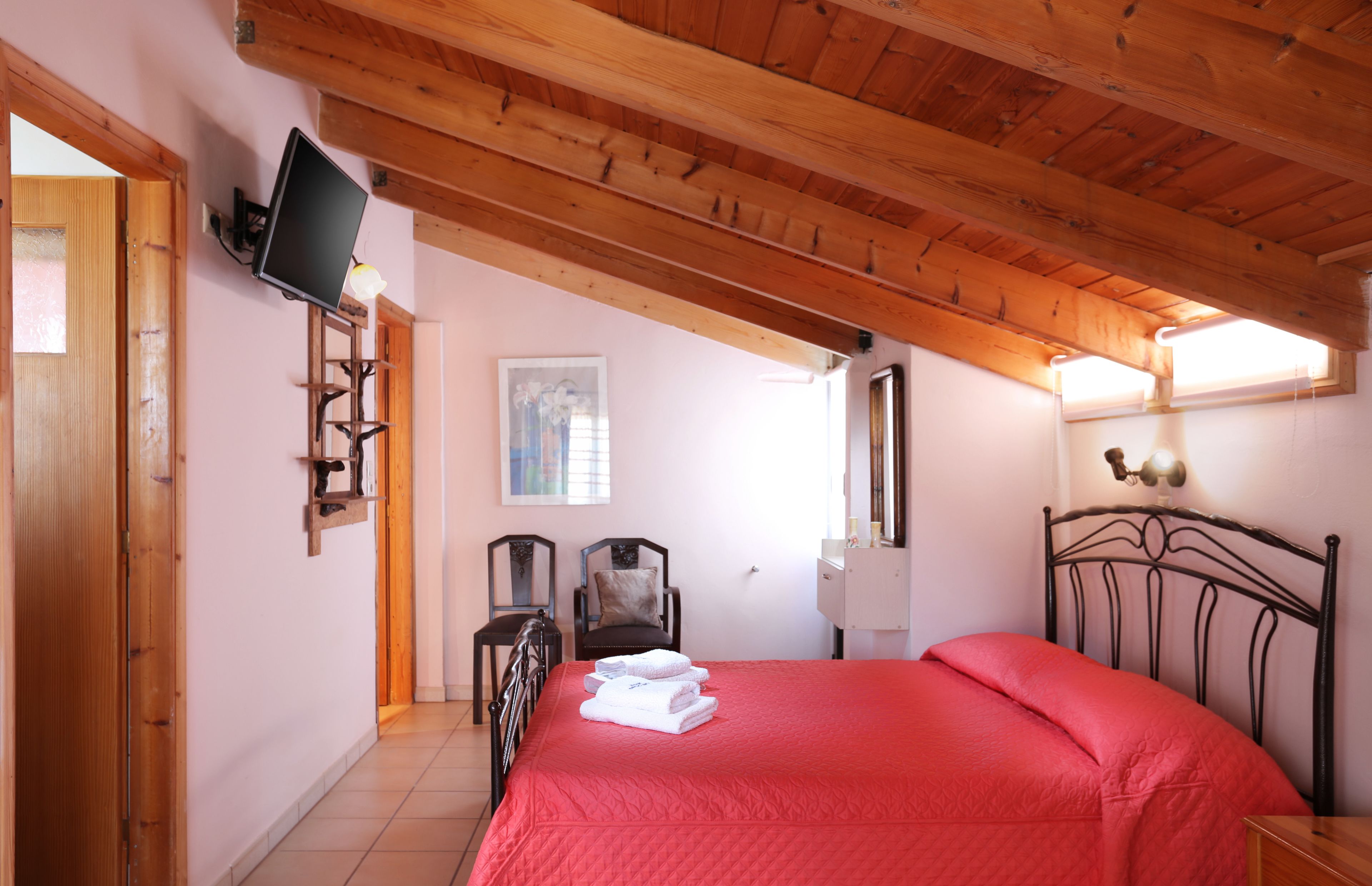 Un dormitorio con colcha roja, vigas de techo de madera, televisor montado y un pequeño escritorio con lámpara.