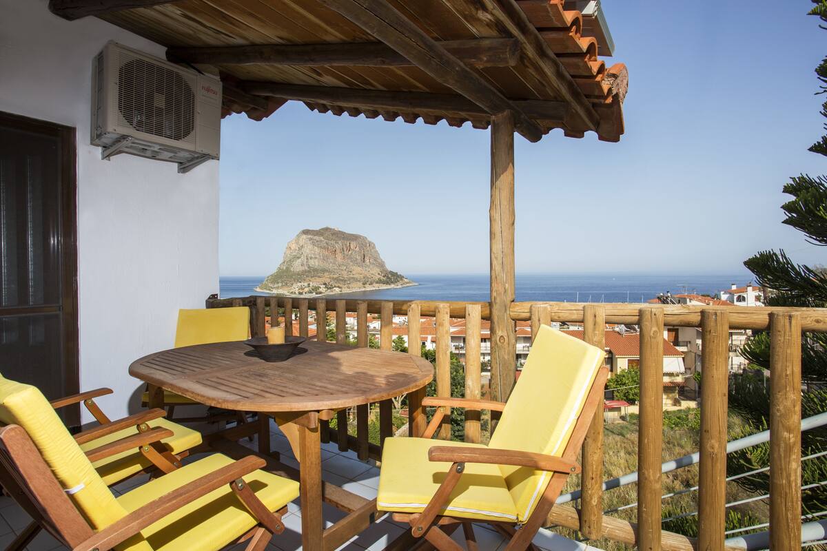 A wooden deck with a round table and yellow chairs, overlooking a coastal view with a prominent rock formation.