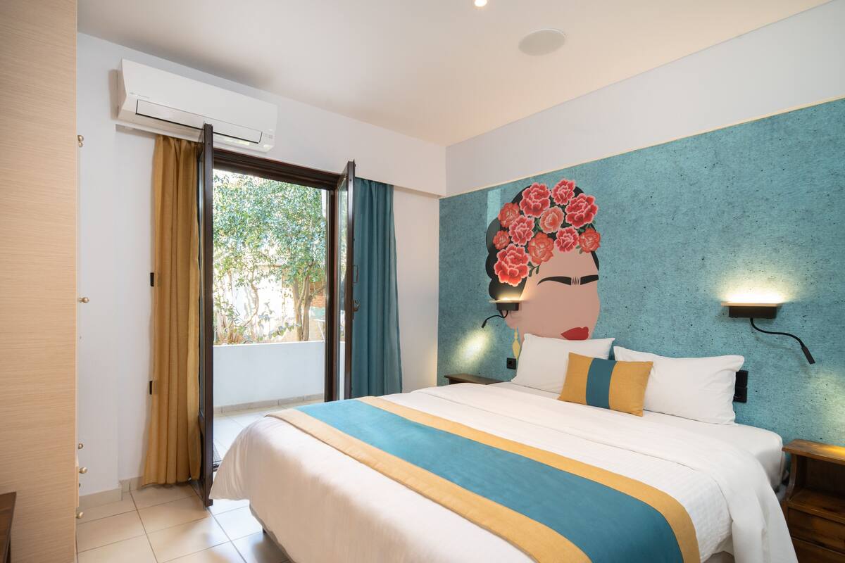 A hotel room with a bed, a window with curtains, an air conditioning unit, and a wall with a floral mural.