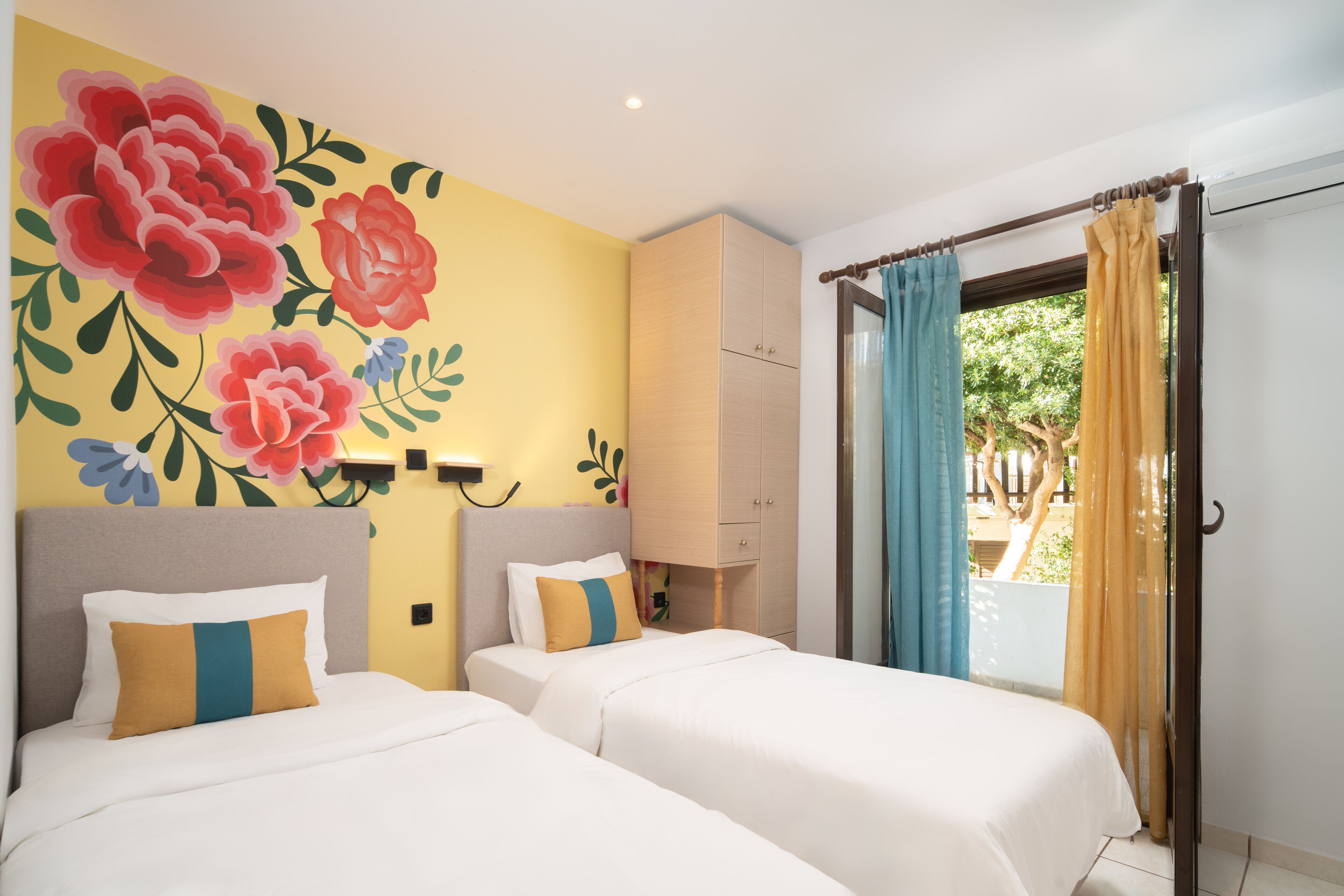 Habitación de hotel con dos camas, un gran mural floral en la pared y vistas al exterior por una puerta corredera.
