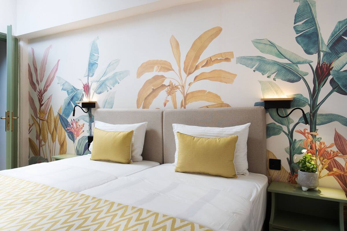 A double bed with white and yellow bedding, two bedside tables with lamps, and a wall with tropical plant murals.