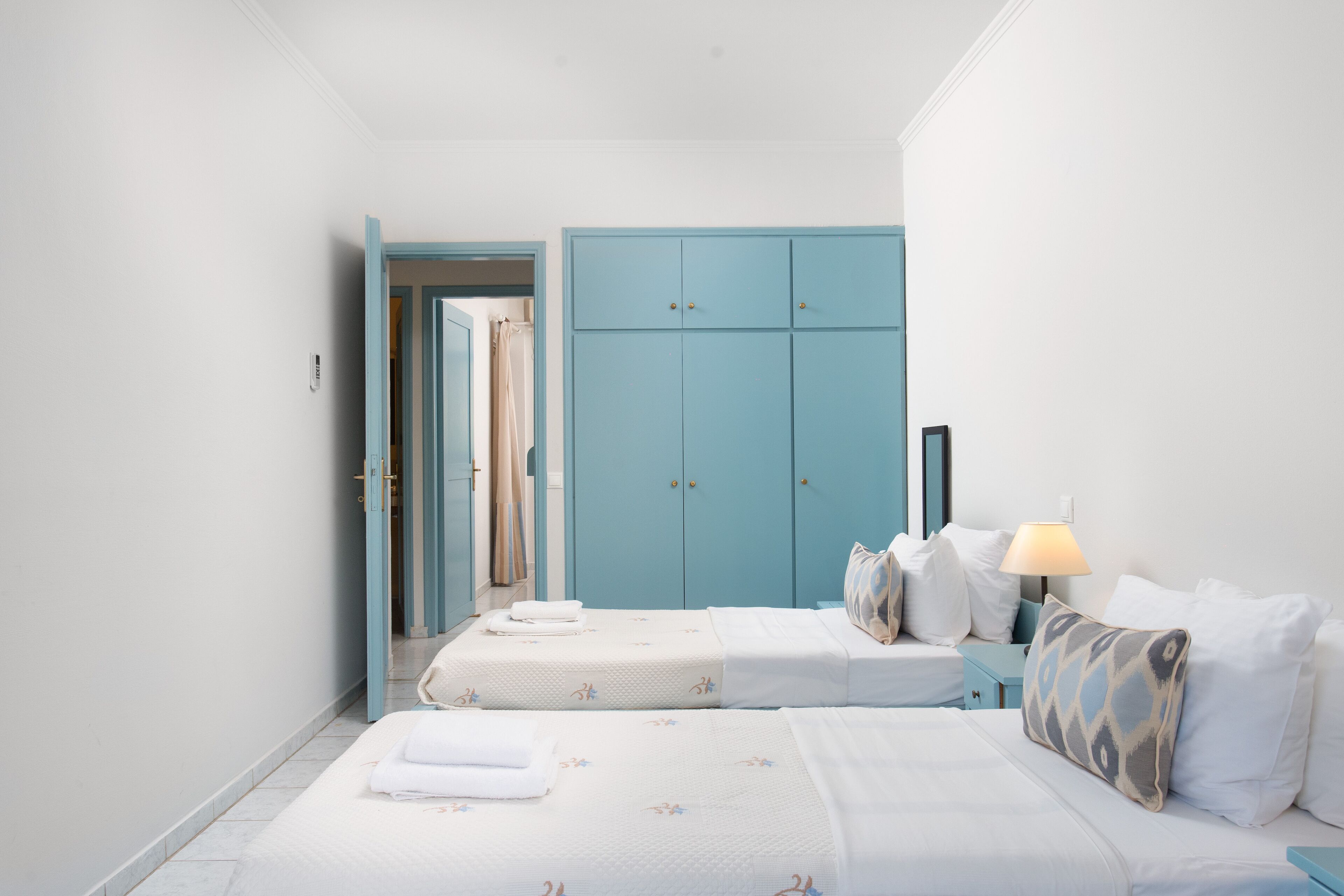 Habitación de hotel con dos camas, un armario azul, una mesita de noche con lámpara y un baño visible a través de una puerta abierta.