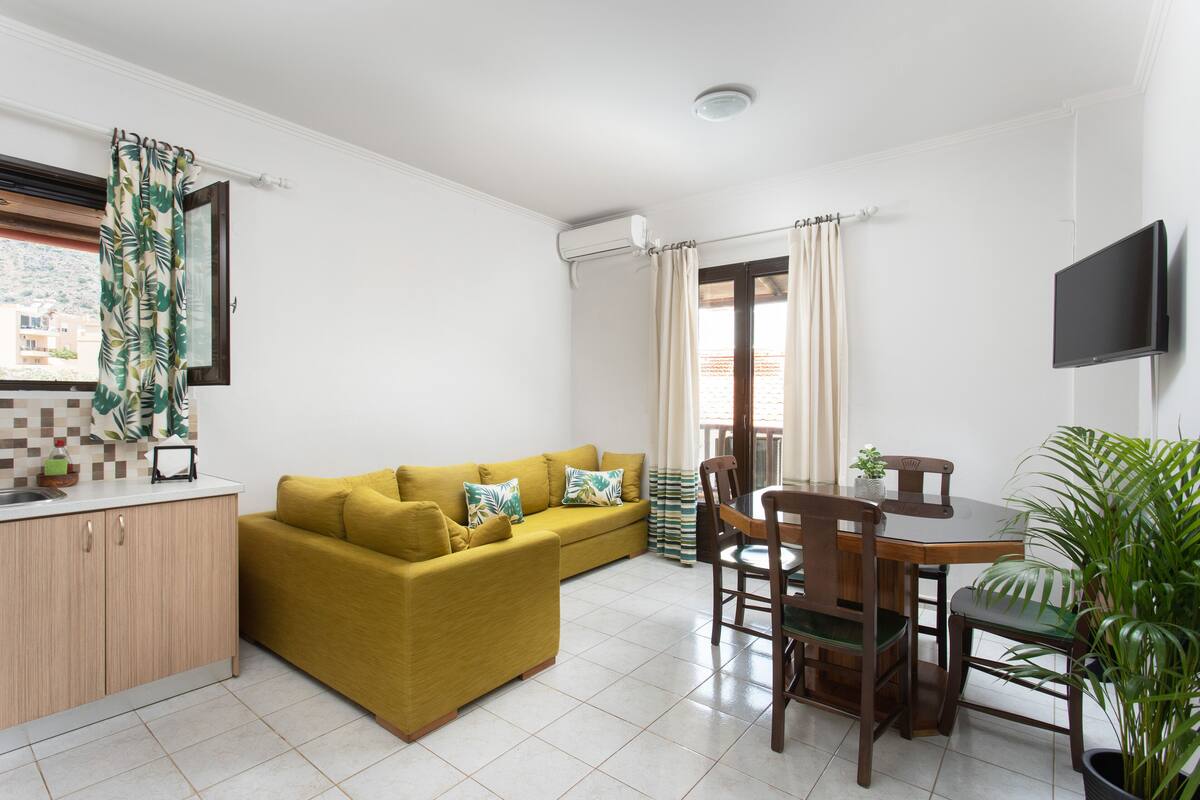 A compact living space with a sofa, dining table, chairs, a small kitchen area, and a TV mounted on the wall.