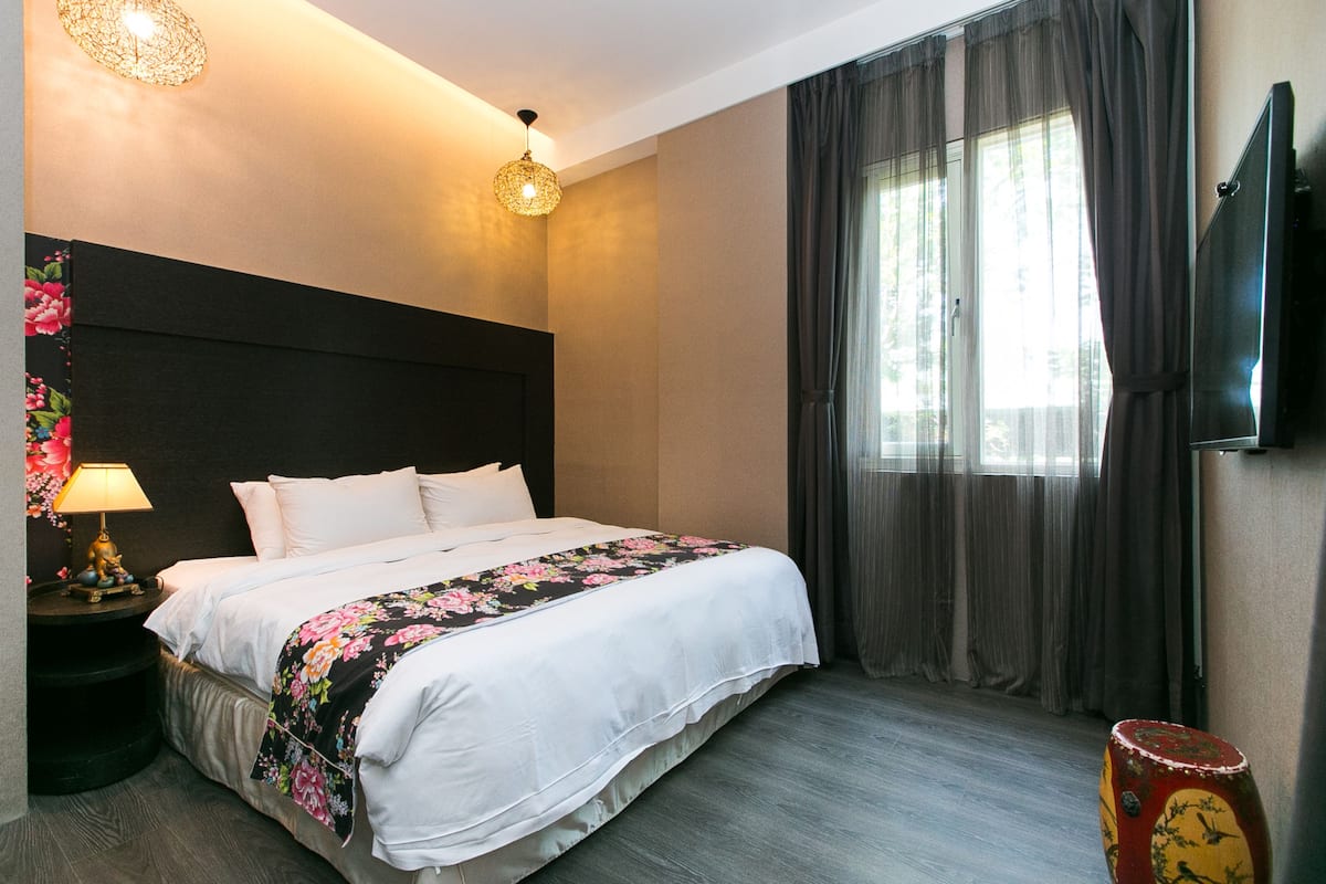 A hotel room with a large bed, a nightstand with a lamp, a television on the wall, and a window with curtains.