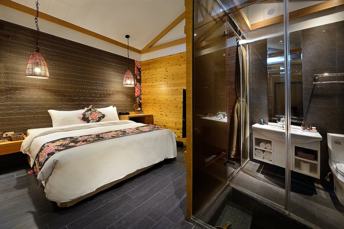 A modern hotel room with a large bed, a wooden accent wall, a glass shower enclosure, and a built-in vanity.