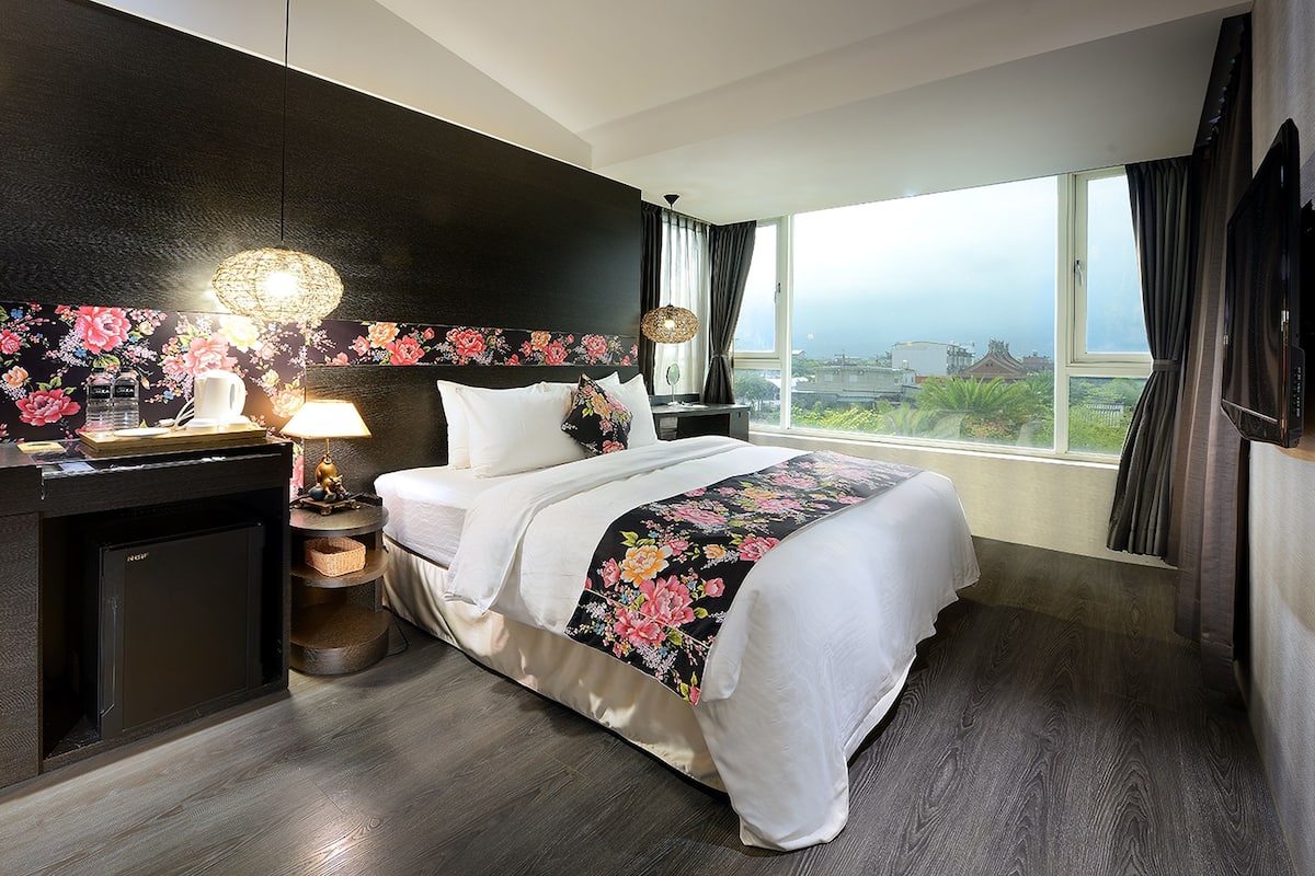 A hotel room with a large bed, a desk with a lamp, a small refrigerator, and a view of the city through a large window.