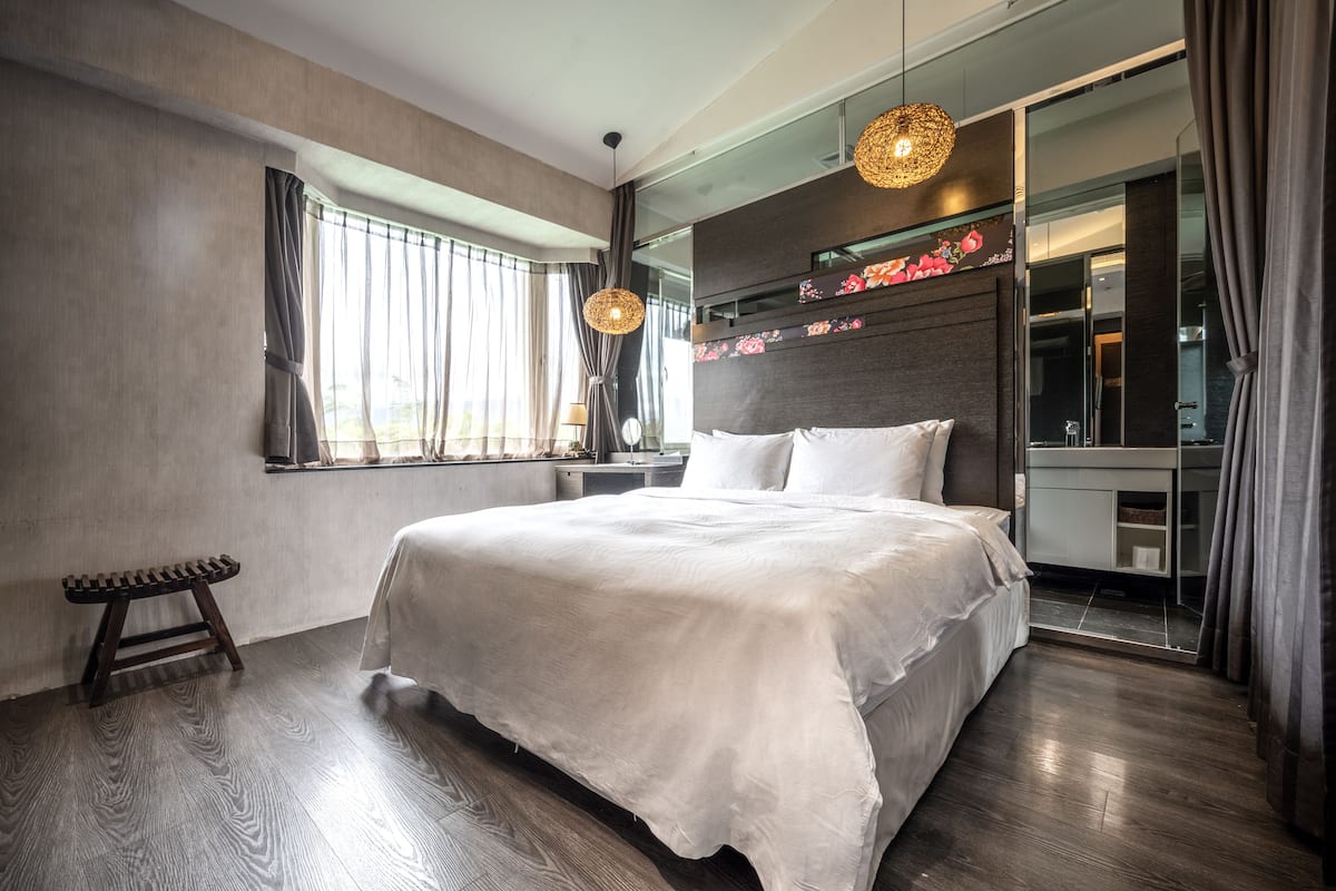 A modern hotel room with a large bed, a small stool, a vanity with a mirror, and a large window with curtains.