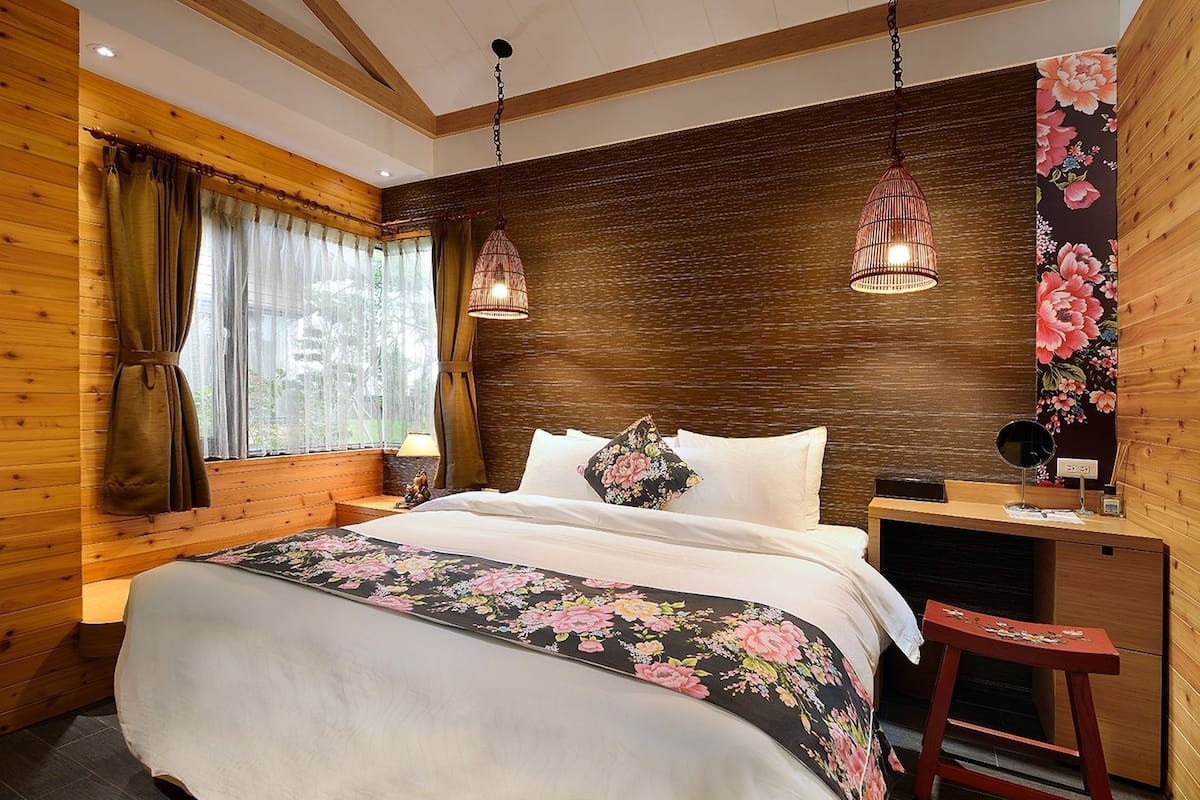 A wooden-paneled room with a bed, two hanging lamps, a nightstand, and a floral wall tapestry.