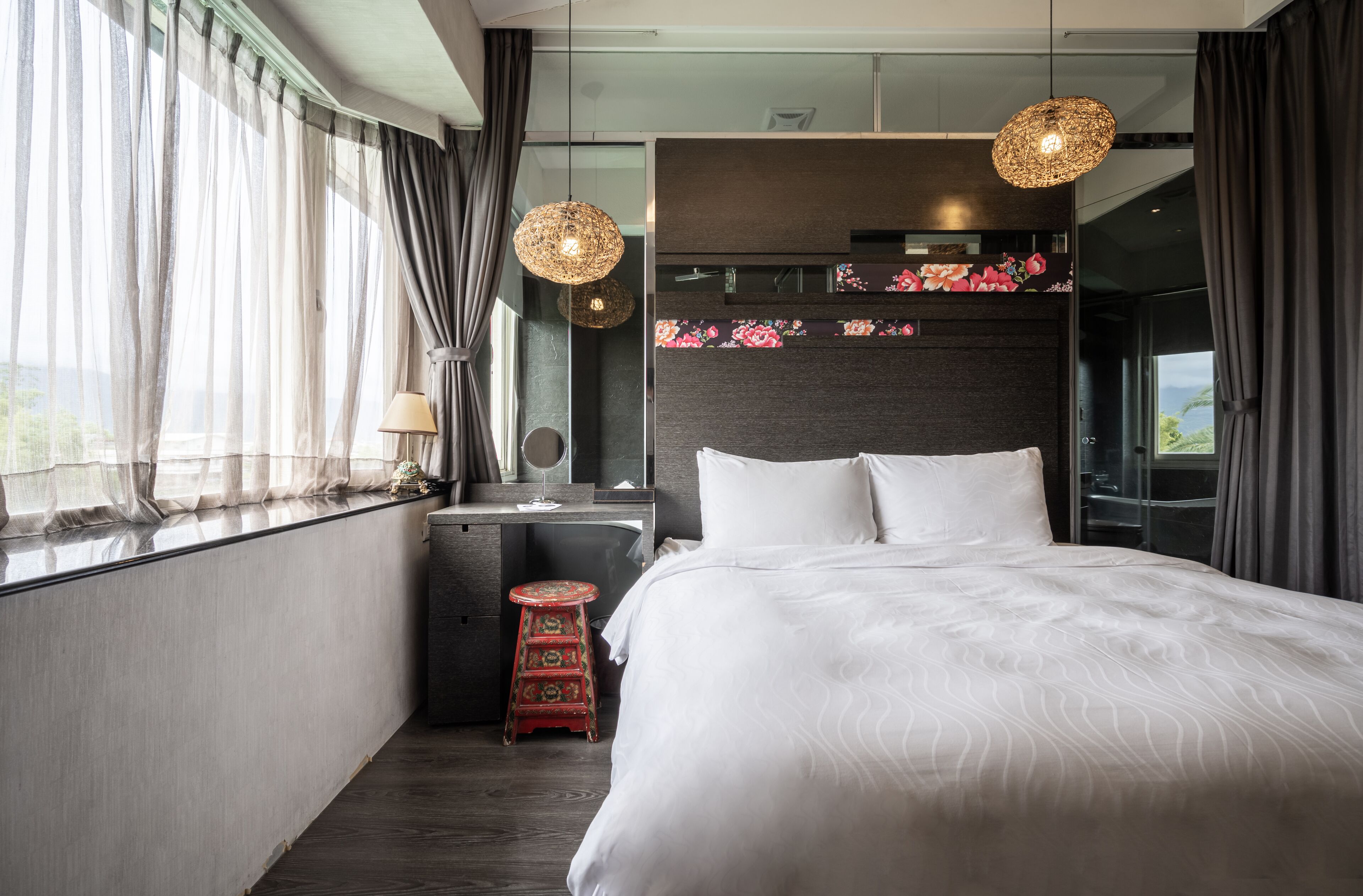 Una habitación de hotel moderna con una cama grande, un escritorio con lámpara y un taburete pequeño. Se ve una ventana con cortinas y vistas al exterior.