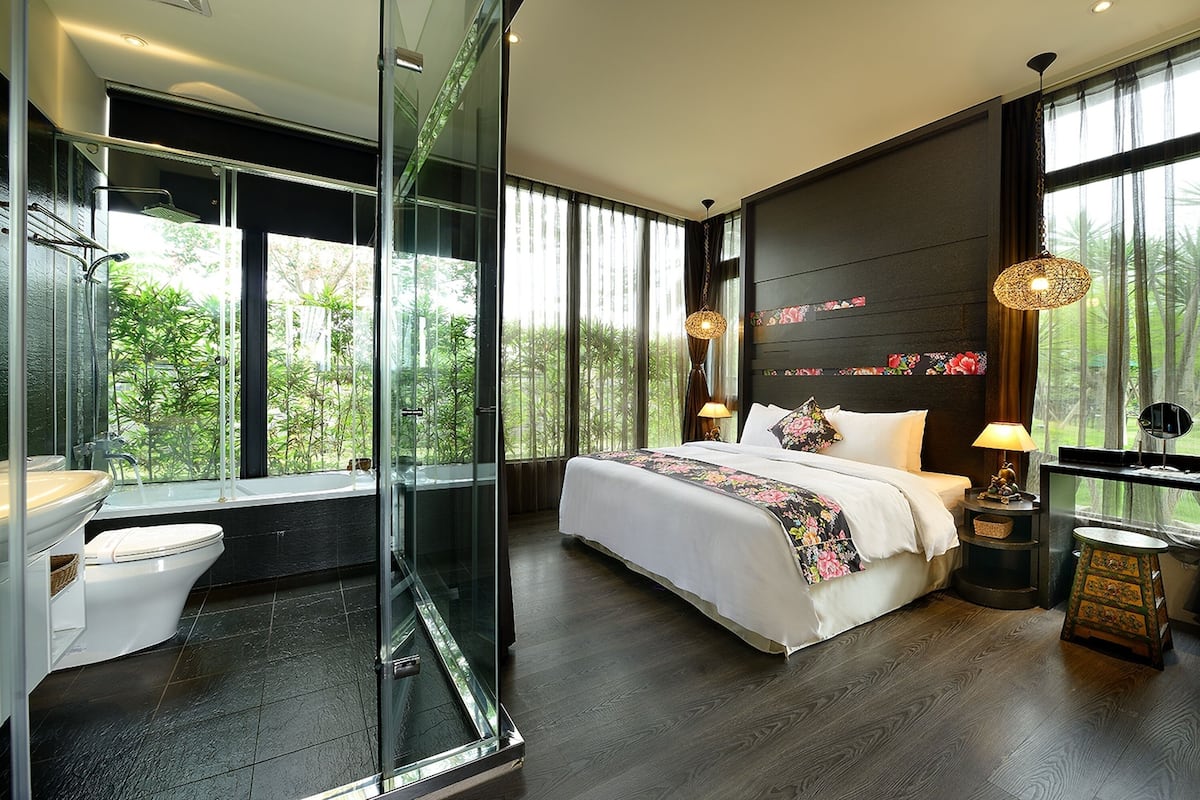 A modern bedroom with a large bed, a glass shower enclosure, and a view of greenery outside.
