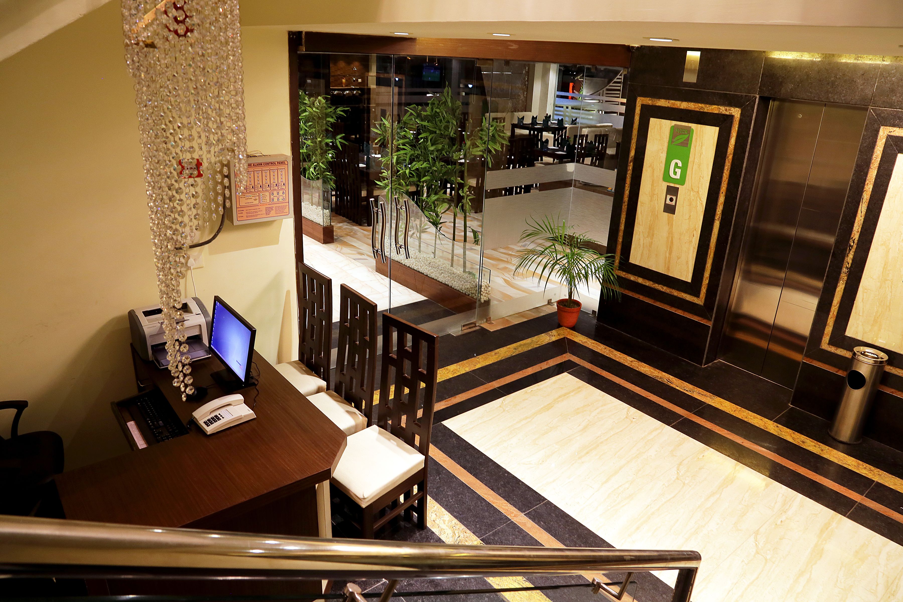 A hotel lobby with a reception desk, chairs, a computer, a printer, a potted plant, and an elevator.