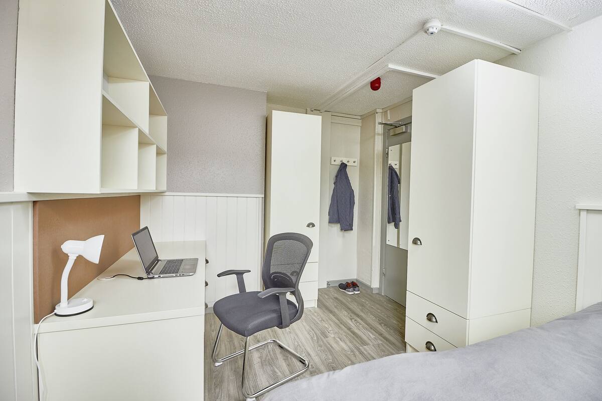 Ein kleines Schlafzimmer mit einem Bett, einem Schreibtisch mit Laptop und Schreibtischlampe, einem Stuhl und einem Kleiderschrank.
