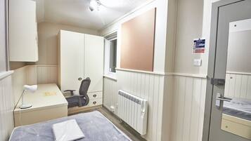 A compact hotel room with a bed, a desk with a chair, a radiator, and a wardrobe.