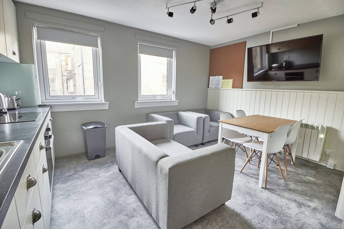 Eine moderne Küche mit einem grauen Sofa, einem Esstisch mit Stühlen und einem an der Wand befestigten Fernseher.
