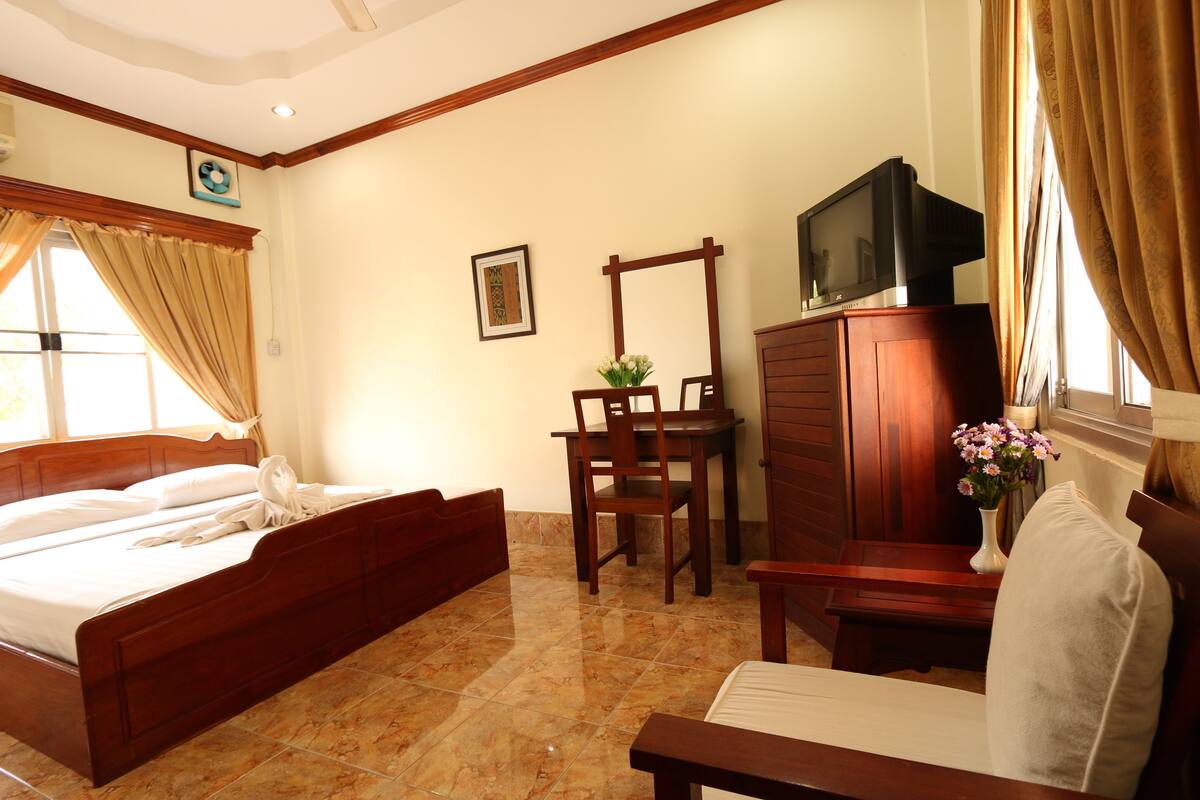 A hotel room with a wooden bed, a wooden desk, a television on a cabinet, and a window with curtains.