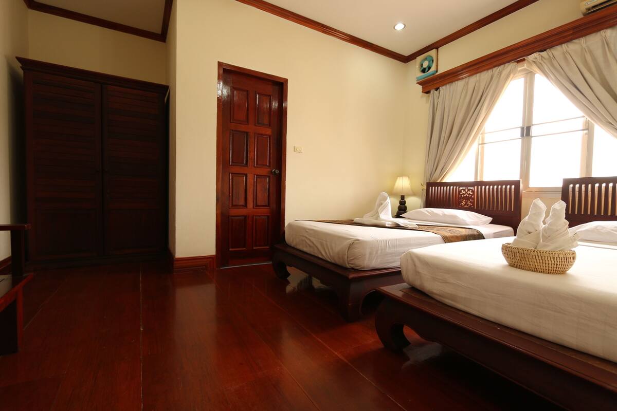 A hotel room with two single beds, wooden flooring, a large wardrobe, and a window with curtains.