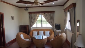 Una habitación con ventilador de techo, televisor sobre una mesita, muebles de mimbre y una ventana con cortinas.