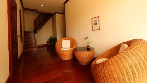 Un pasillo con suelo de madera, sillas de mimbre, una pequeña mesa con una planta y una escalera al fondo.