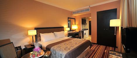 Ein Hotelzimmer mit einem großen Bett, Nachttischlampen, einem Schreibtisch inklusive Lampe, einem Sessel, einem Fernseher und einem Fenster mit Vorhängen.
