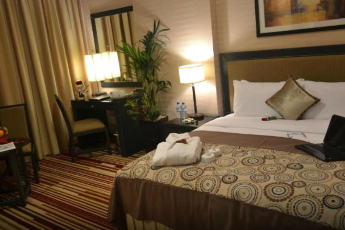 Une chambre d’hôtel avec un lit, un bureau, une chaise, une lampe, une plante et un tableau au mur.