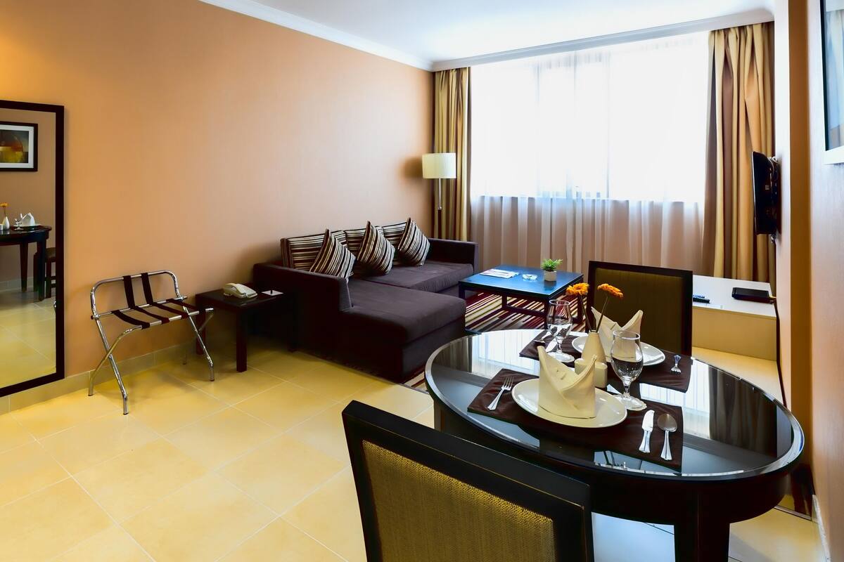 A hotel room with a sofa, a dining table set for two, a wheelchair, a mirror, and a window with curtains.