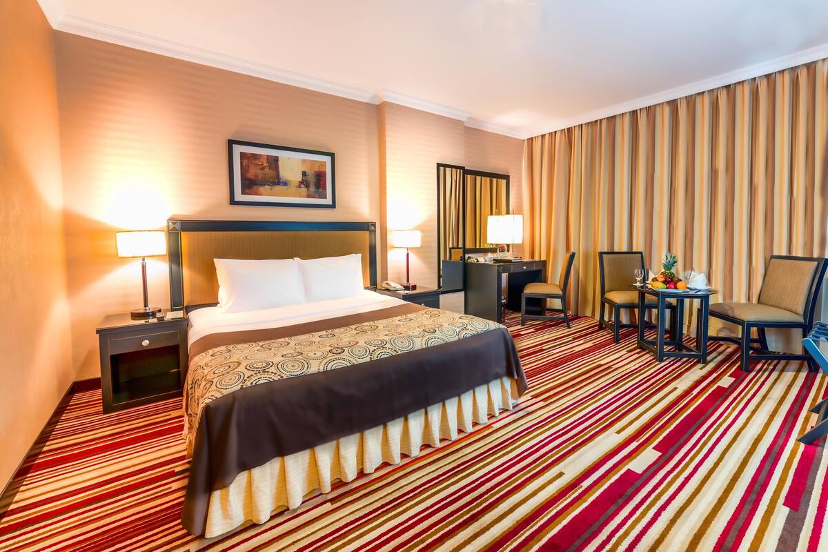 A hotel room with a large bed, two bedside tables with lamps, a desk with a chair, and a large window with curtains.