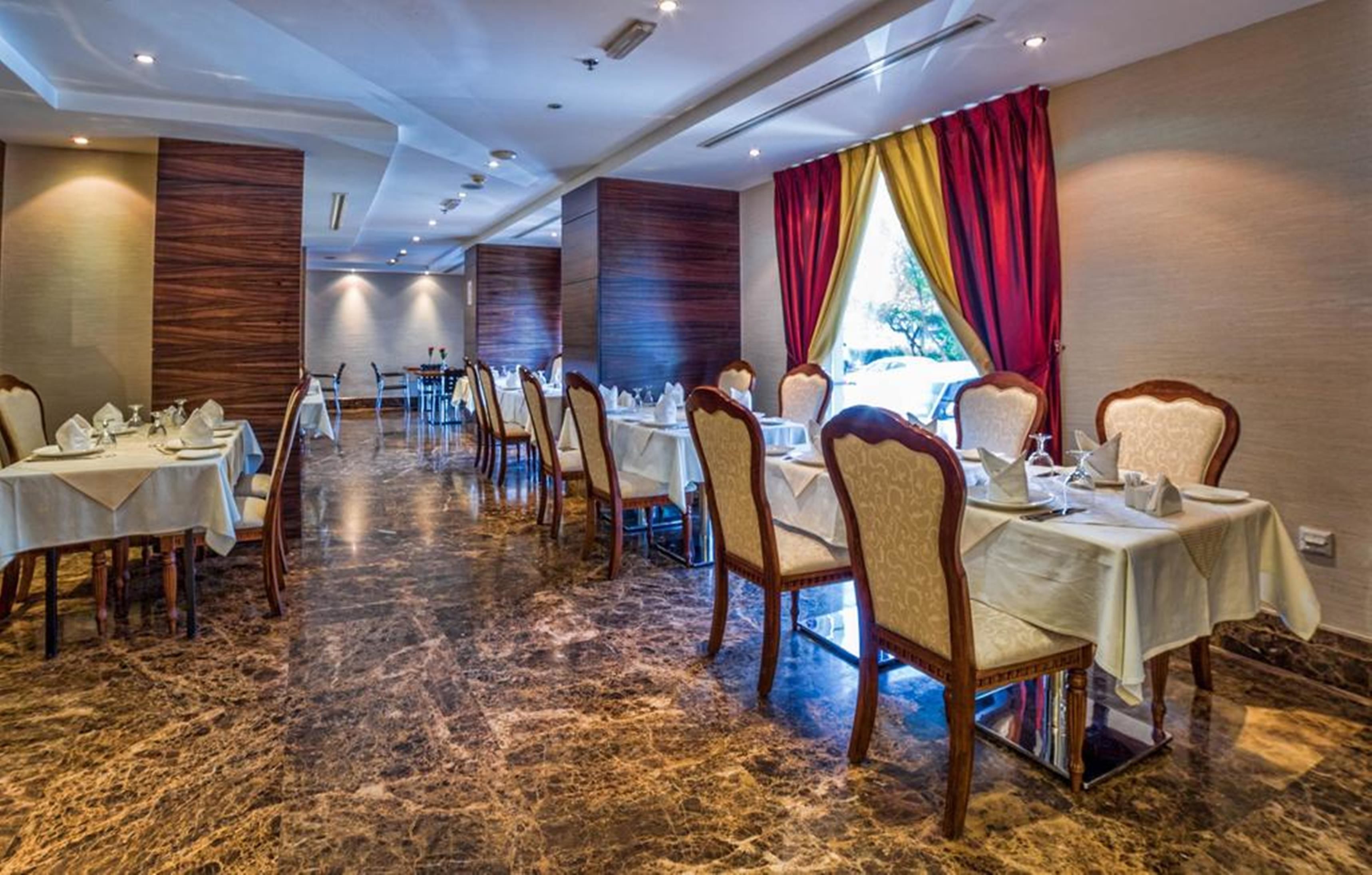 A hotel dining area with marble flooring, rows of tables with chairs, and a view of the outdoors through a window with red curtains.