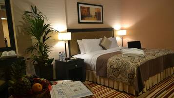 A hotel room with a bed, two bedside lamps, a potted plant, a fruit bowl, and a newspaper on the nightstand.