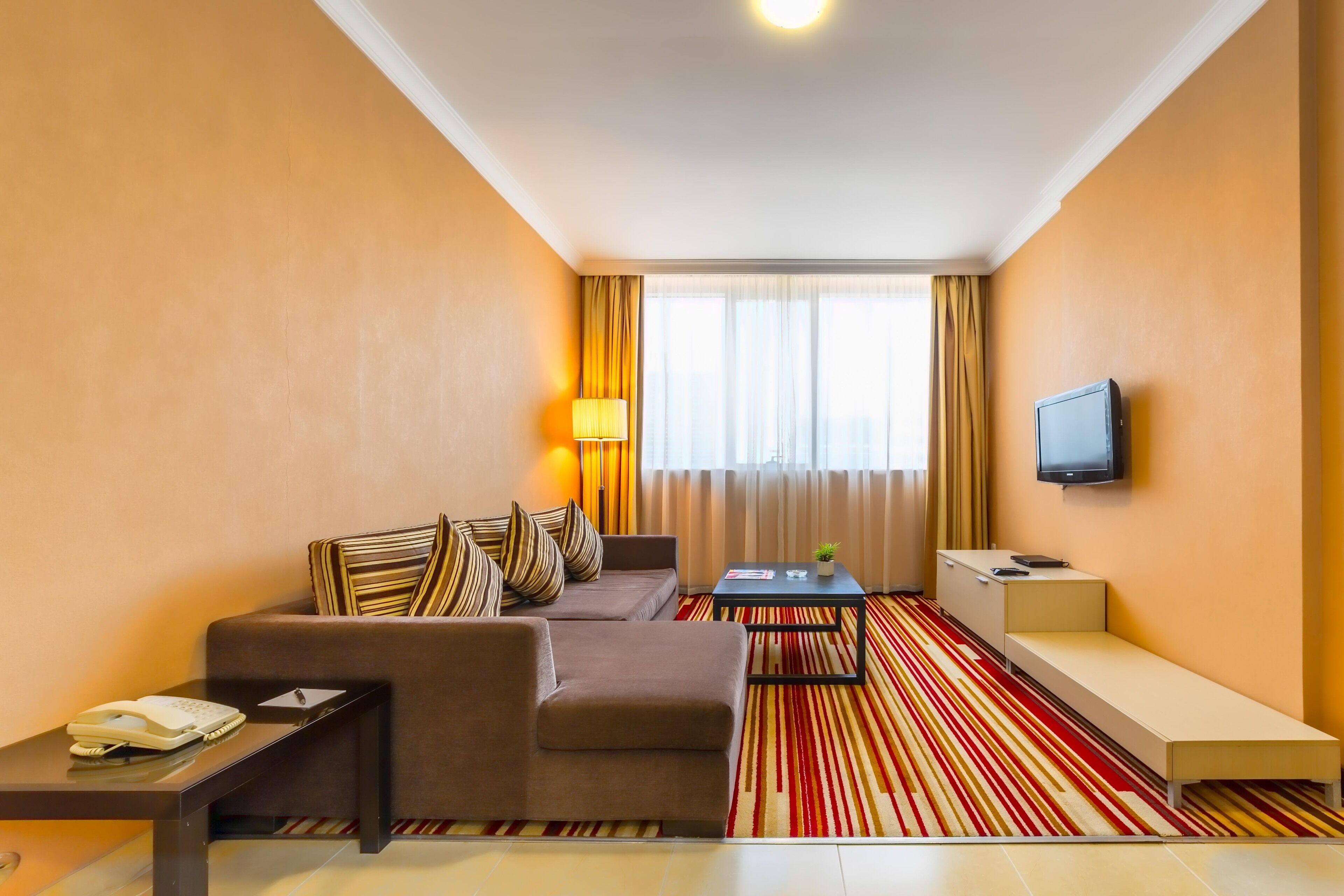 A hotel room with a sofa, a small table with a lamp, a TV mounted on the wall, and a striped carpet.