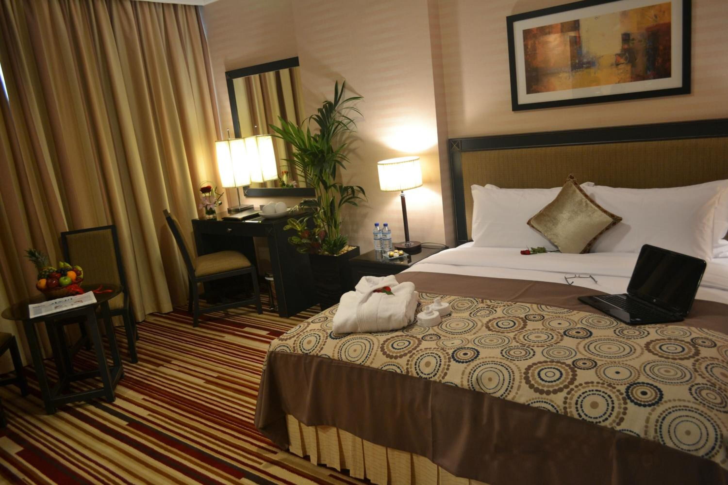 A hotel room with a bed, a desk, a chair, a lamp, a laptop, and a plant.