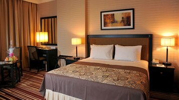Habitación de hotel con una cama grande, dos lámparas de noche, un escritorio con una silla, una lámpara de escritorio, un cuadro en la pared y una alfombra estampada.
