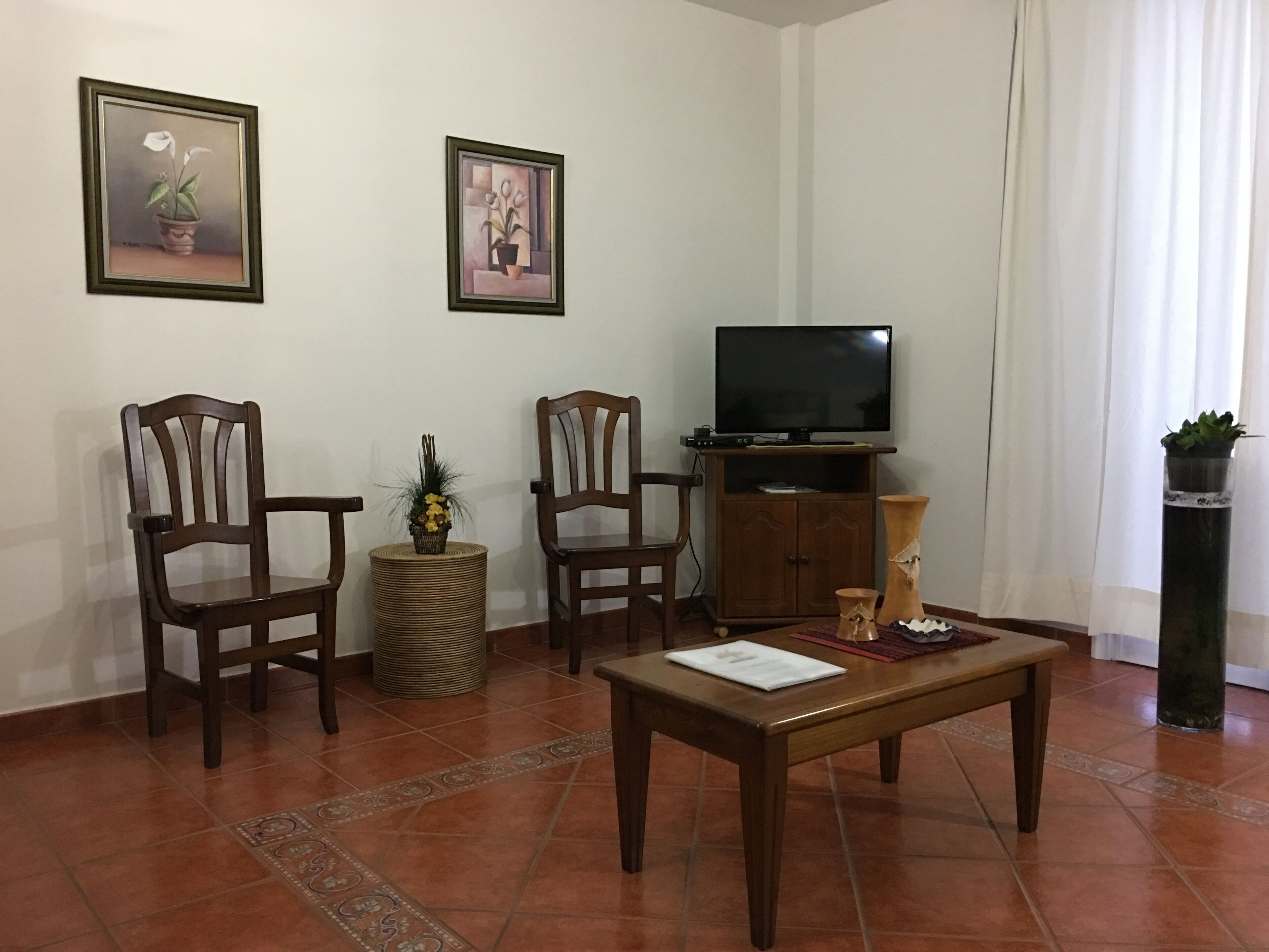 Een woonkamer met twee houten stoelen, een klein rond tafeltje met een document, een televisie op een houten kast en twee ingekaderde foto’s aan de muur.