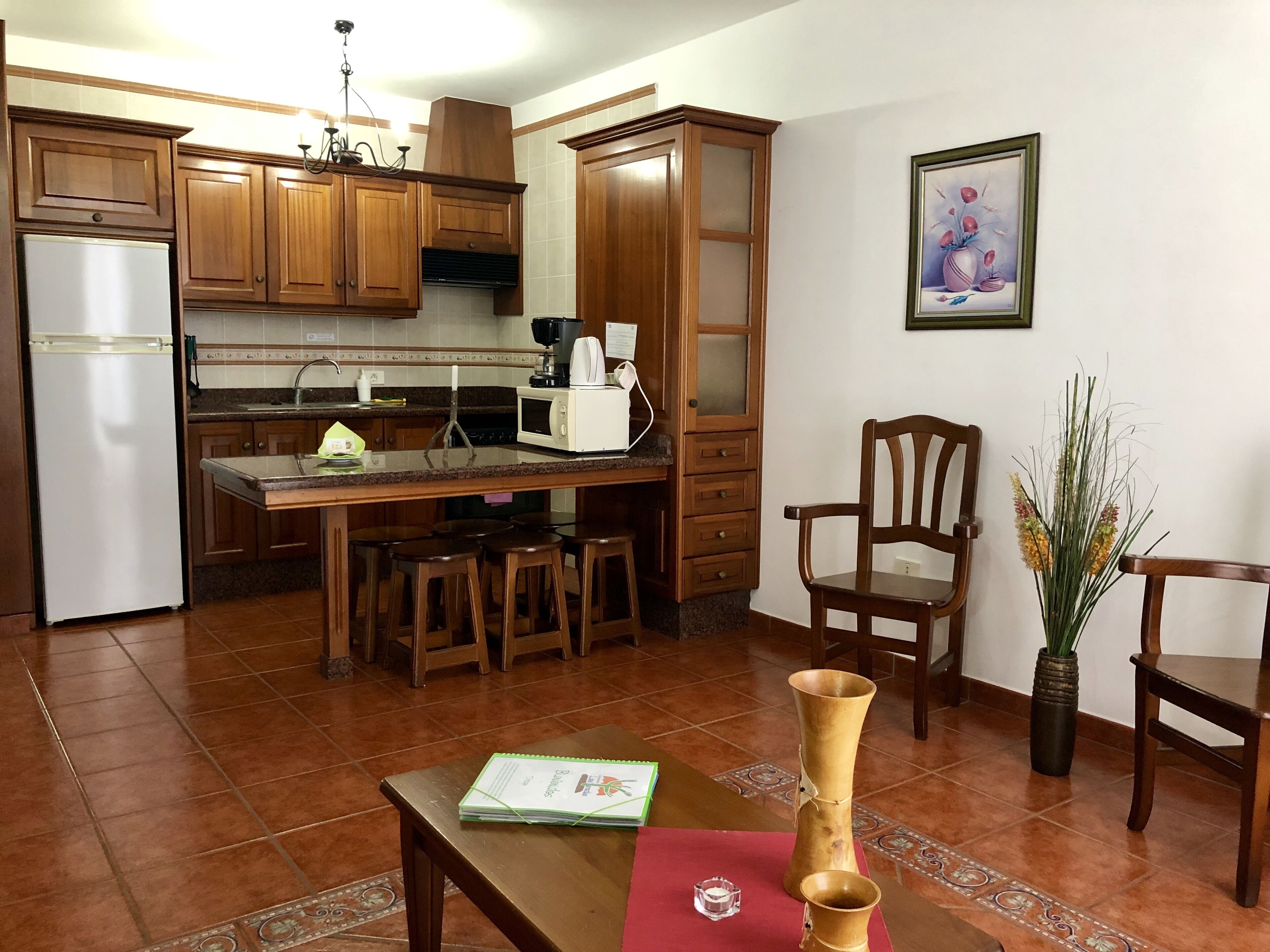 A compact kitchen with wooden cabinets, a refrigerator, a microwave, and a coffee maker. There are two wooden chairs and a table with a book and a vase.