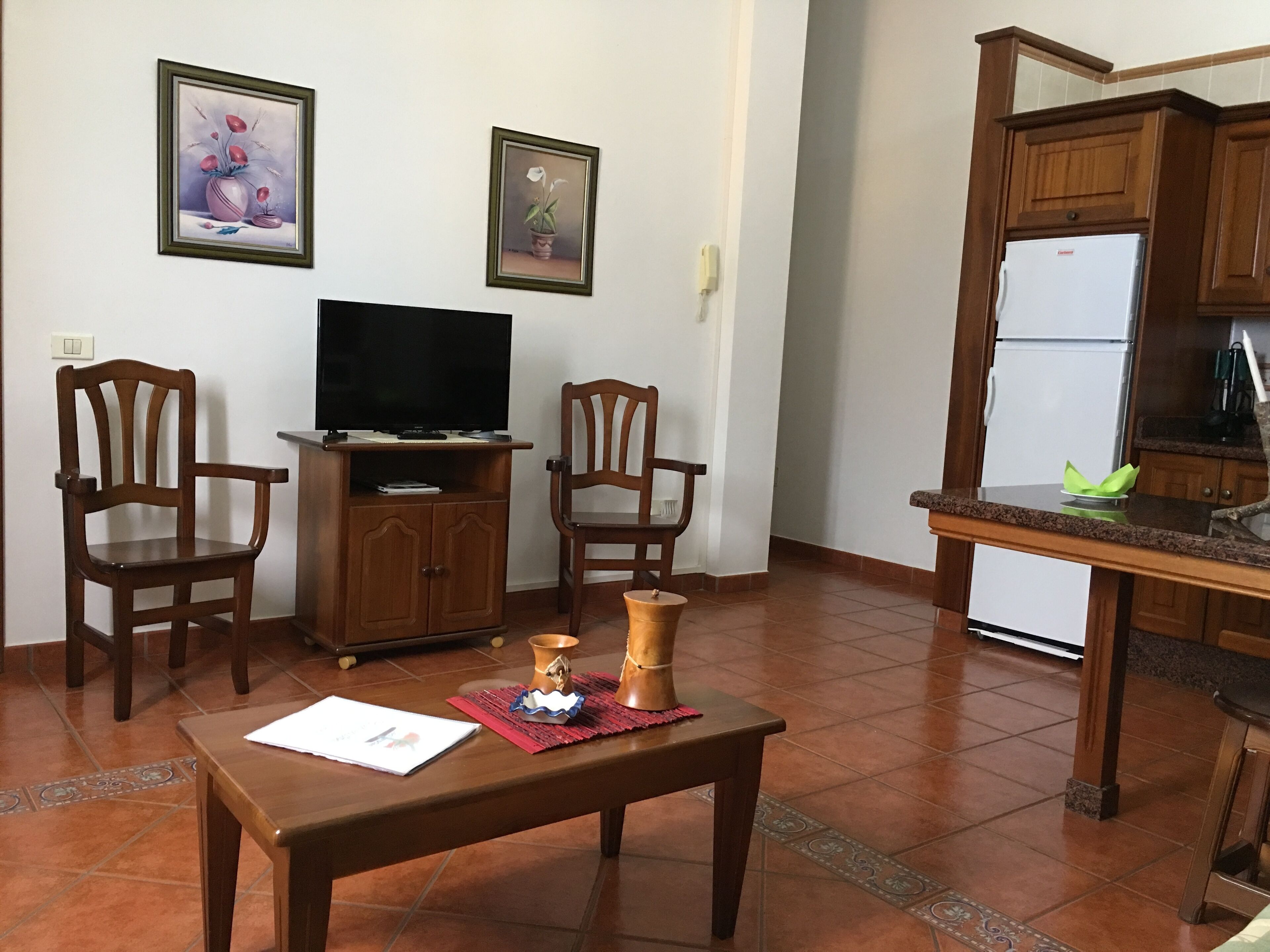 Un salon comprenant un téléviseur, deux fauteuils en bois, une petite table ornée d’un objet décoratif, ainsi qu’un coin cuisine avec un réfrigérateur et des placards.