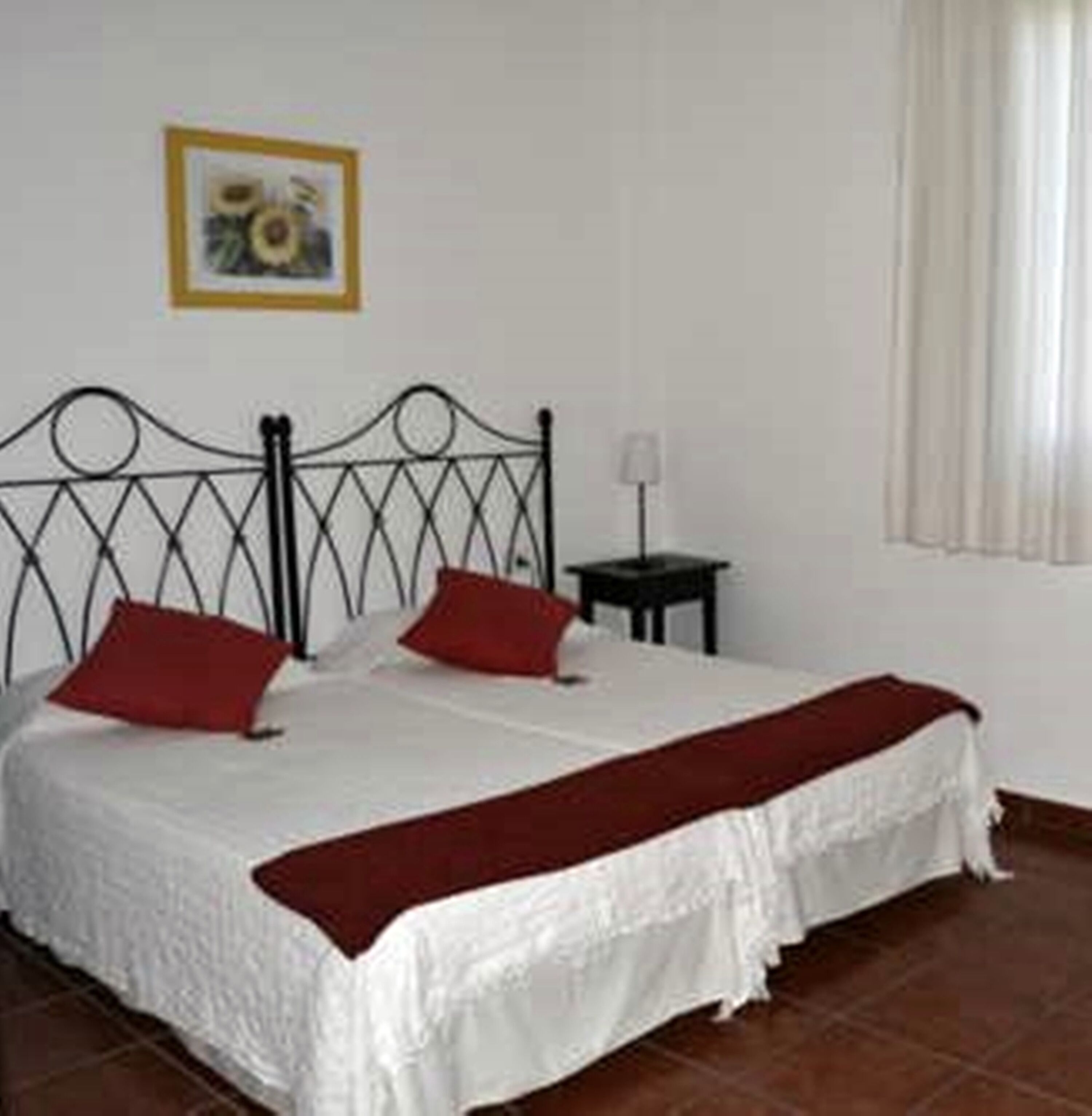 A bedroom with a bed, two red pillows, a white bedspread, a red throw blanket, a small bedside table with a lamp, and a framed picture on the wall.