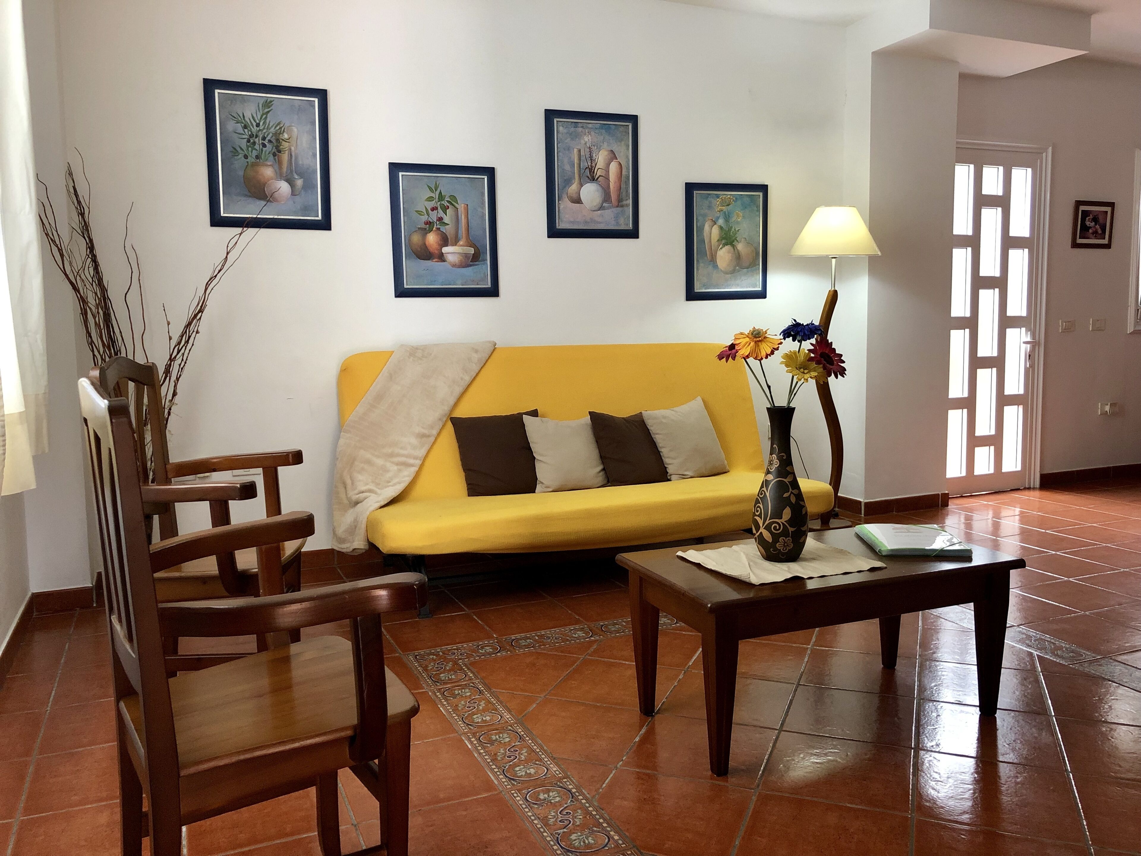 A living room with a yellow sofa, wooden chairs, a coffee table, and a vase with flowers. There are framed paintings on the wall.