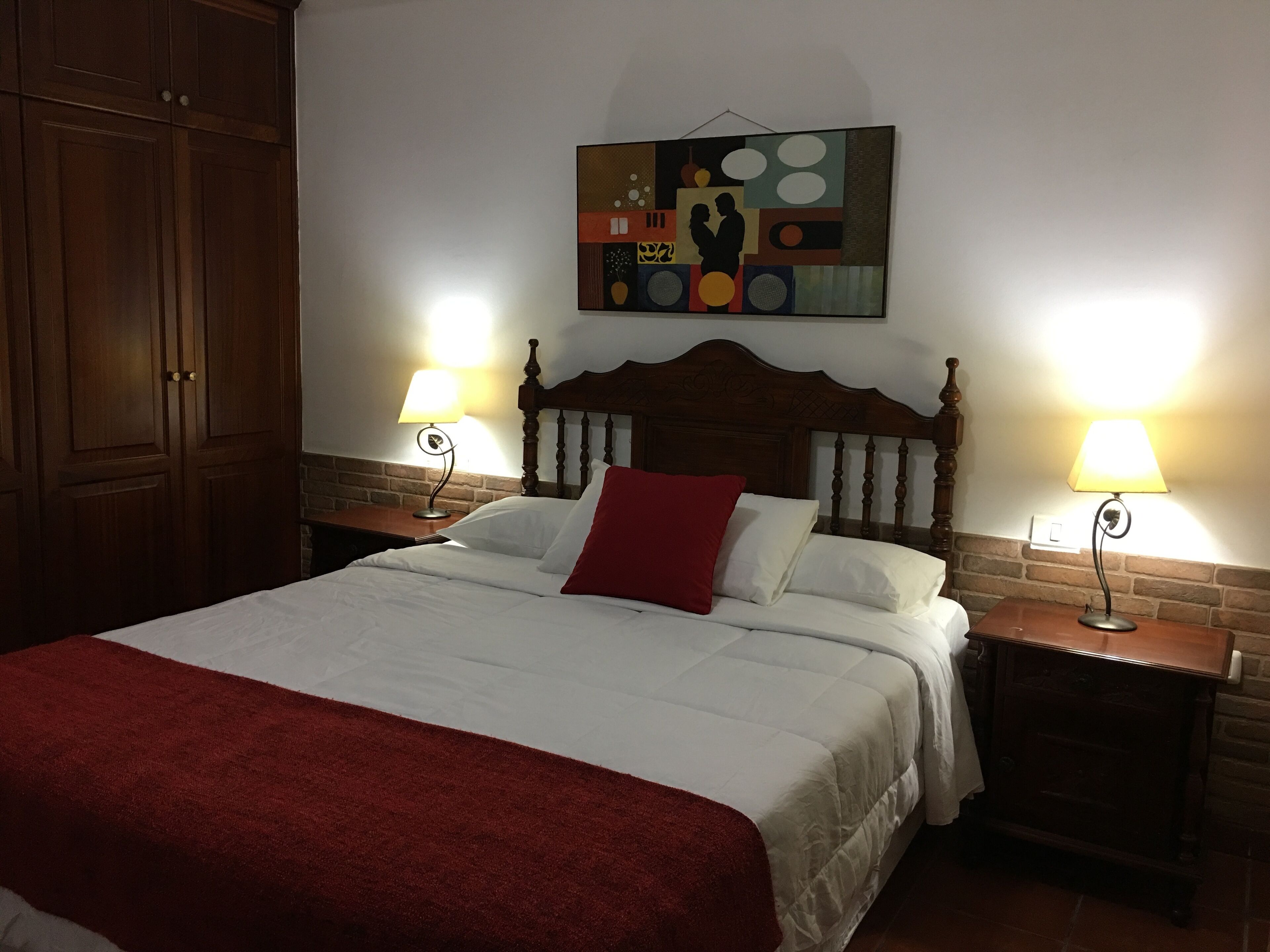 Een netjes opgemaakt bed met een rode dekensprei, twee bedlampjes en een houten hoofdbord.