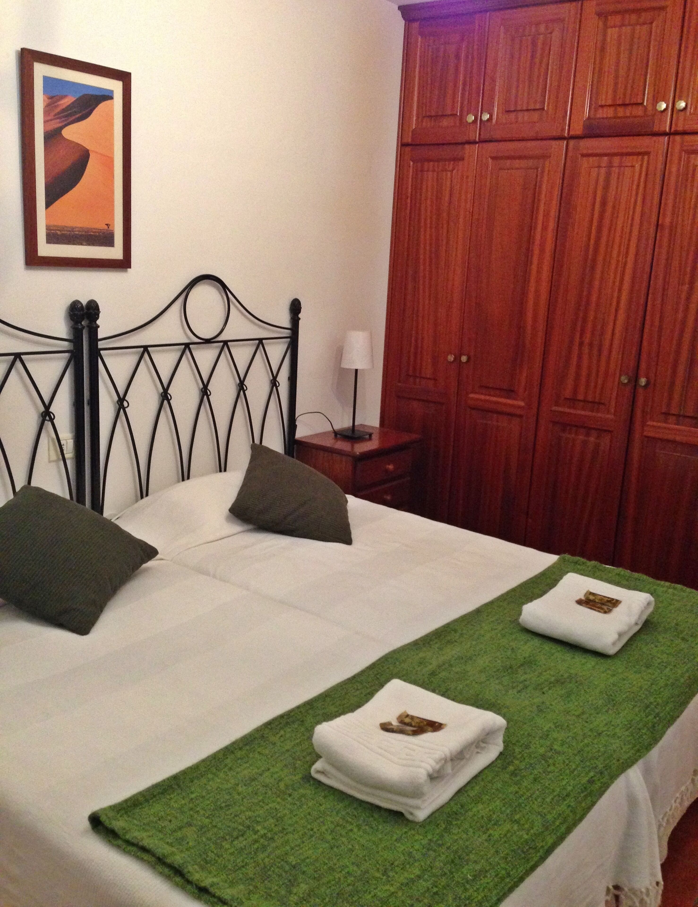 Una cama bien tendida con dos almohadas, una toalla verde y un cuadro en la pared.