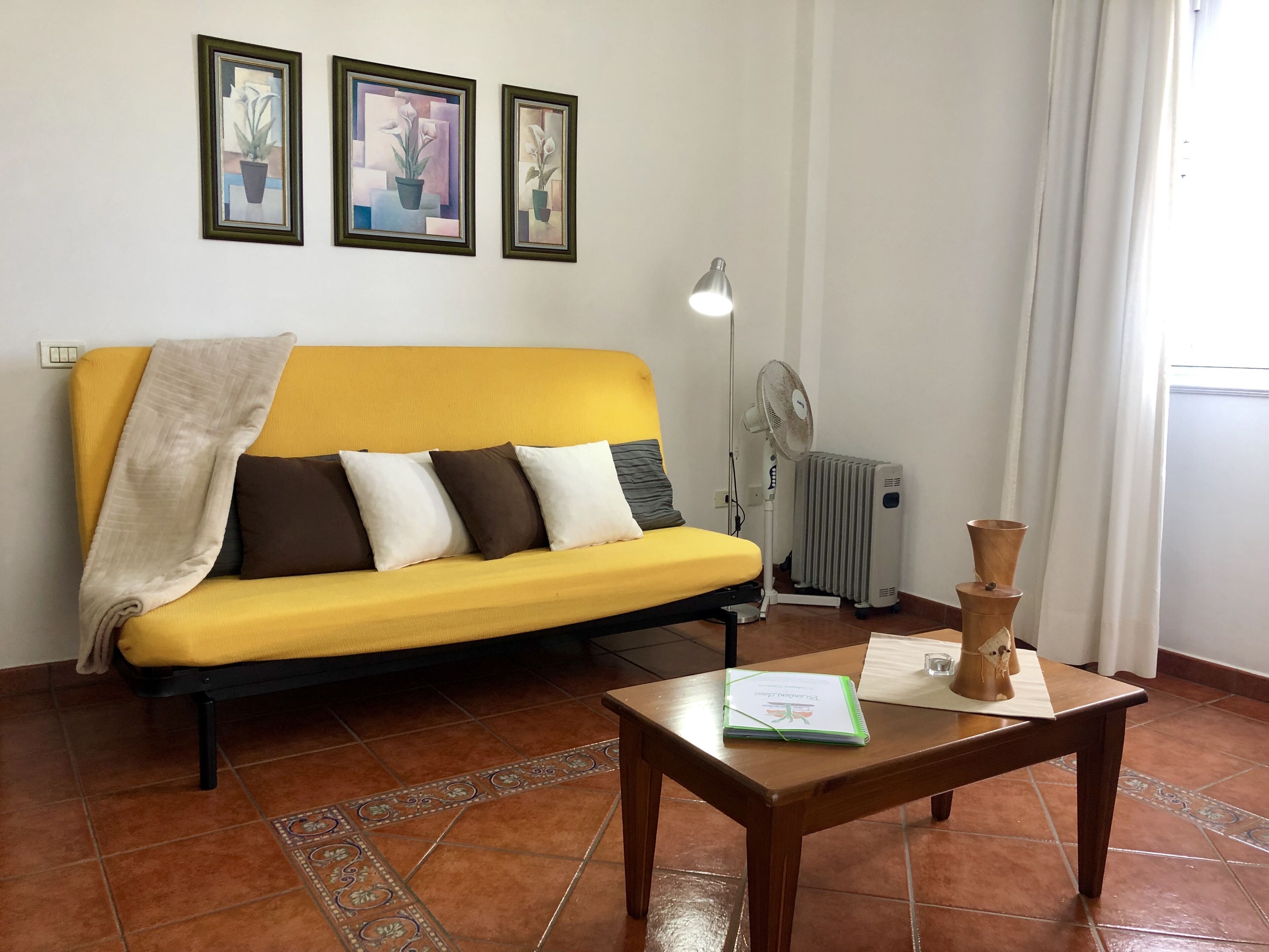 Um sofá amarelo com uma manta, uma mesa de madeira com um caderno, um abajur e um radiador.