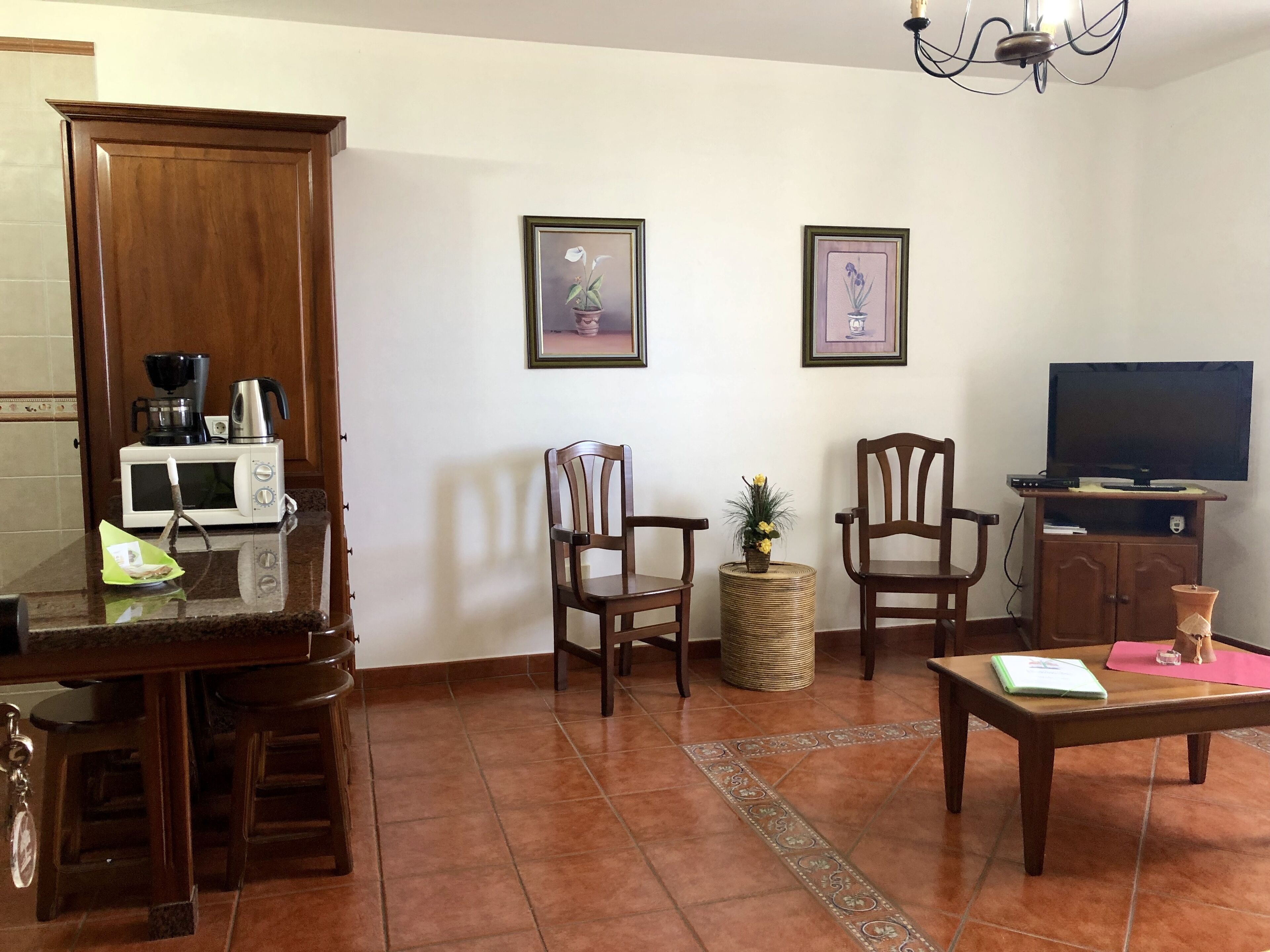 A living room with a wooden cabinet, a microwave, two chairs, a small table, a television, and framed pictures on the wall.