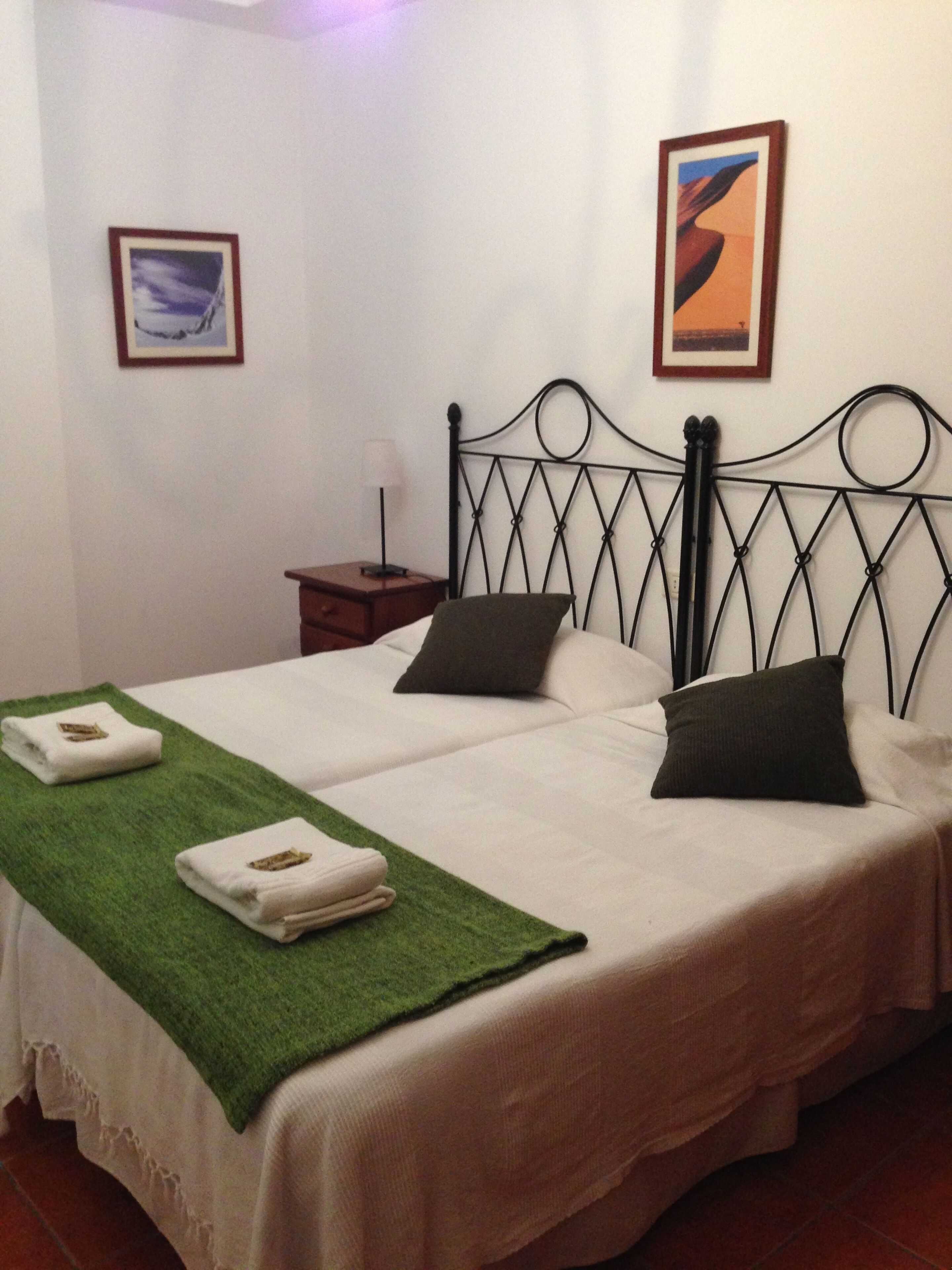 A double bed with a green throw blanket, two pillows, and a nightstand with a lamp.