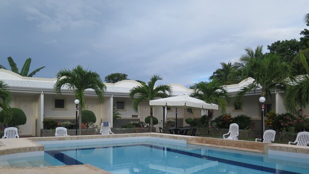 A swimming pool with lounge chairs and umbrellas, surrounded by palm trees and a building with numbered doors. - Villa del Pueblo (Panglao)