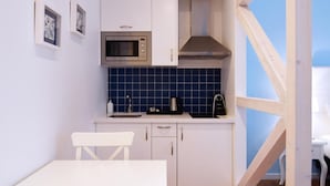 Cocina compacta con armarios blancos, microondas, estufa y fregadero. Hay una mesa de comedor con dos sillas. Las paredes están pintadas de azul, y una ventana con marco de madera.