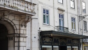 Un edificio histórico con detalles arquitectónicos ornamentados, incluyendo un balcón con barandillas de hierro forjado y un cartel de 'Pratas' en la planta baja.