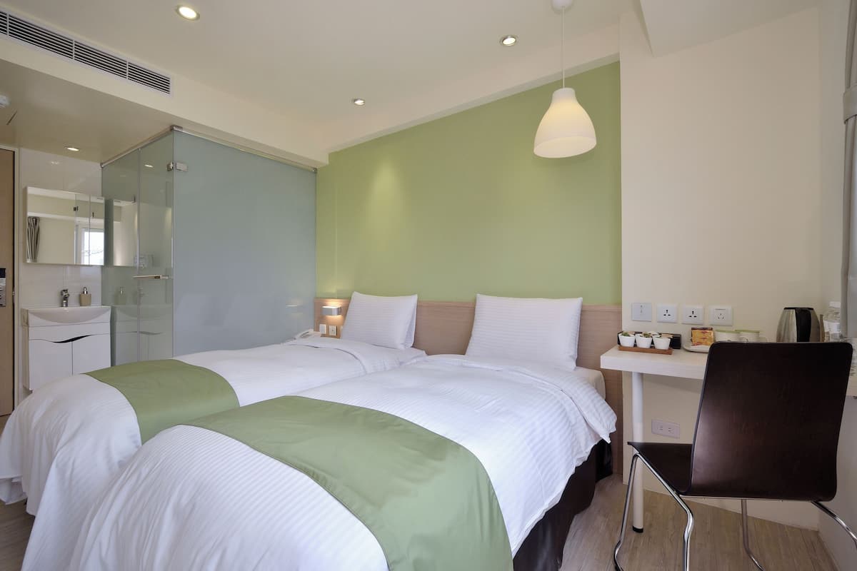 A hotel room with two beds, a bedside table, a chair, a desk with a lamp, and a mirror.