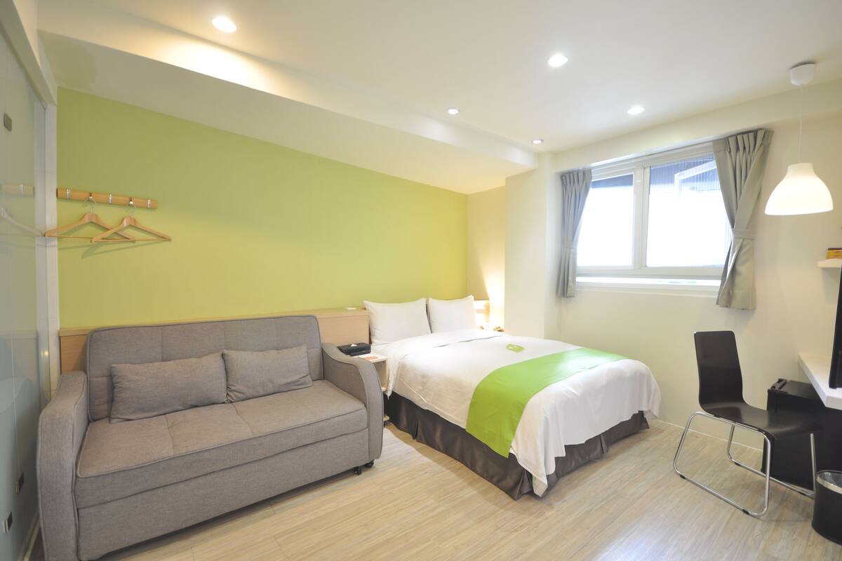 A hotel room with a bed, a grey sofa, a desk, and a window with curtains.