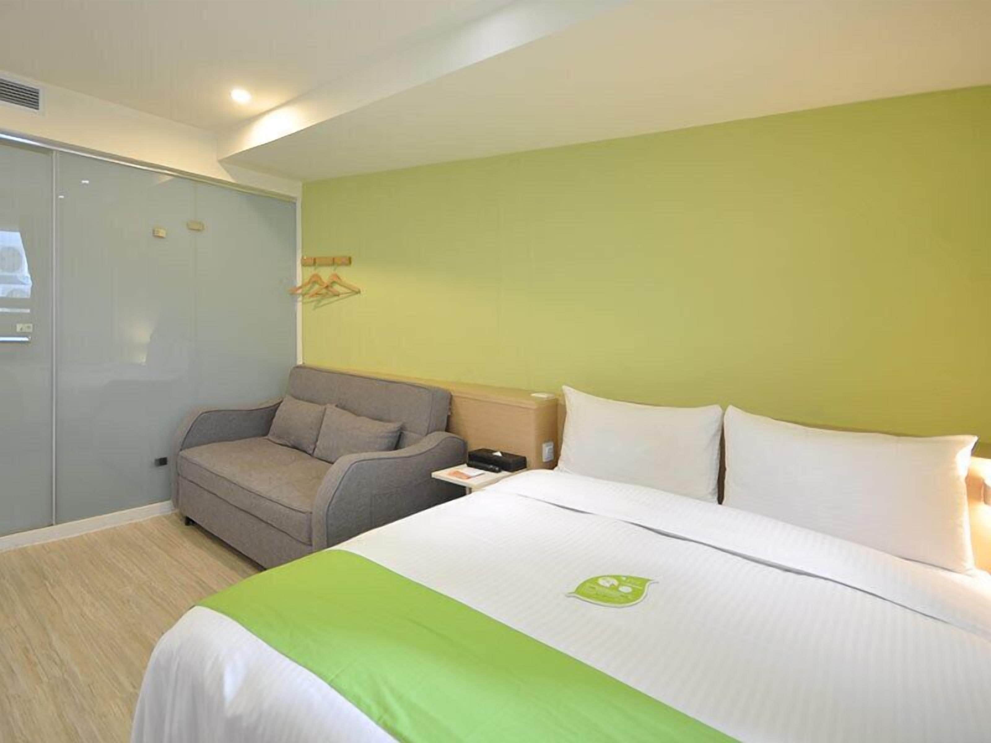 A hotel room with a bed, a grey sofa, a nightstand, and a wardrobe.