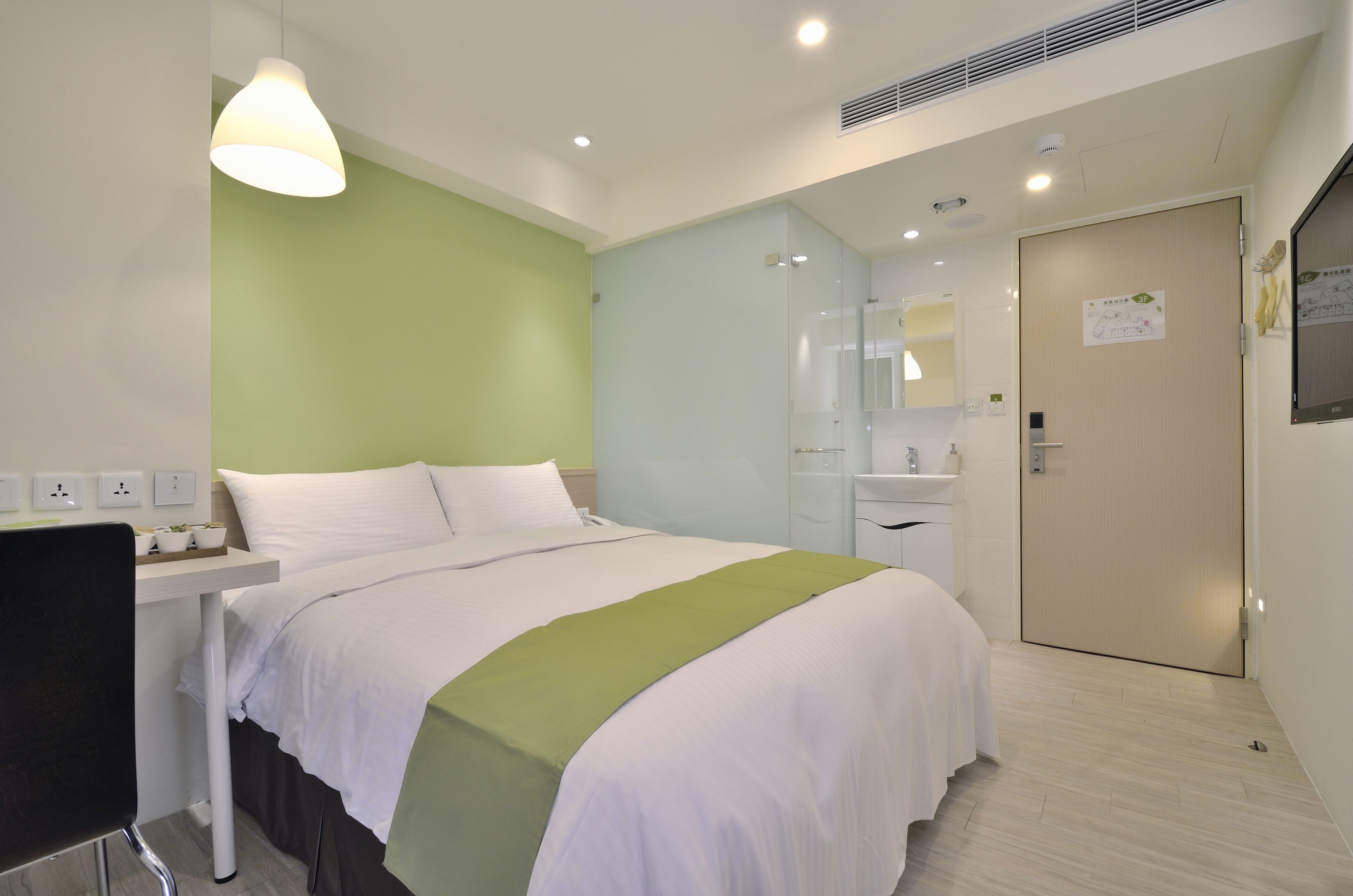 A hotel room with a bed, bedside table, lamp, and a door with a sign.