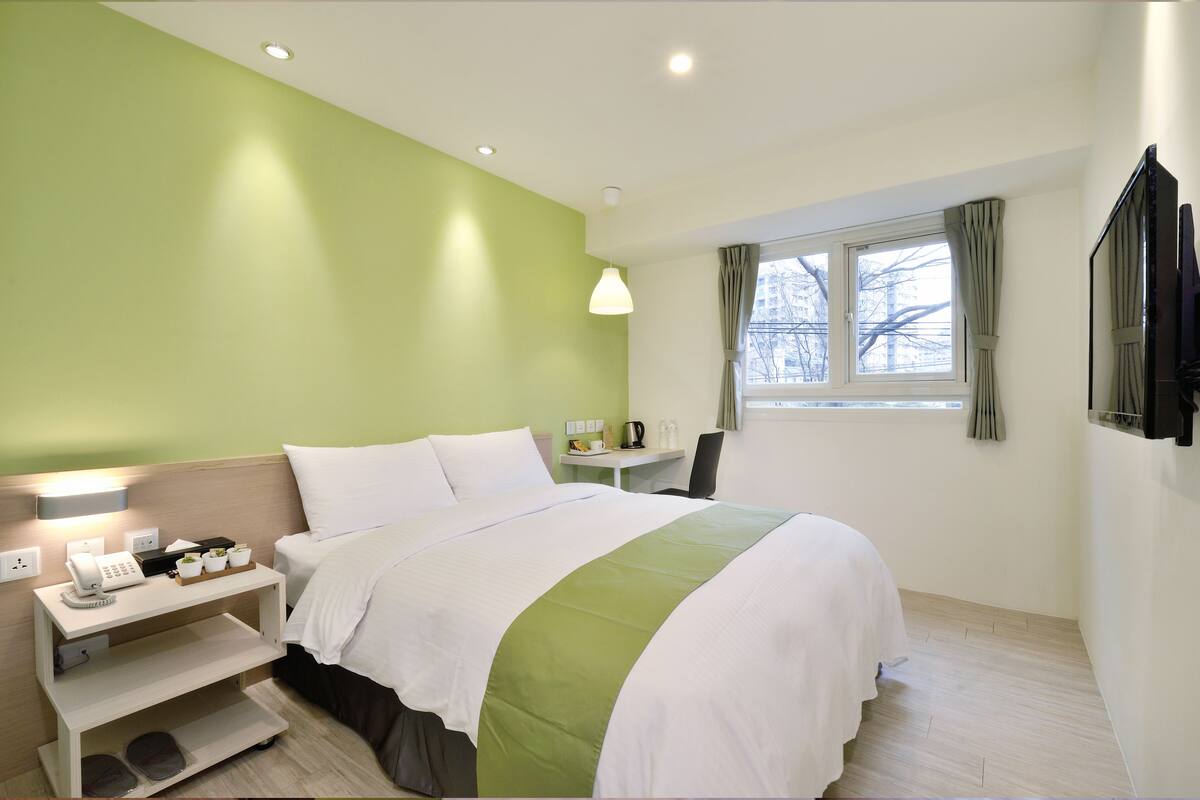 A hotel room with a bed, bedside table, lamp, and a window with curtains.