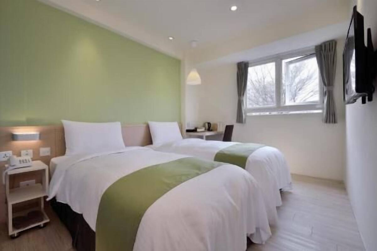 A hotel room with two beds, a desk, a chair, a nightstand, and a window with curtains.